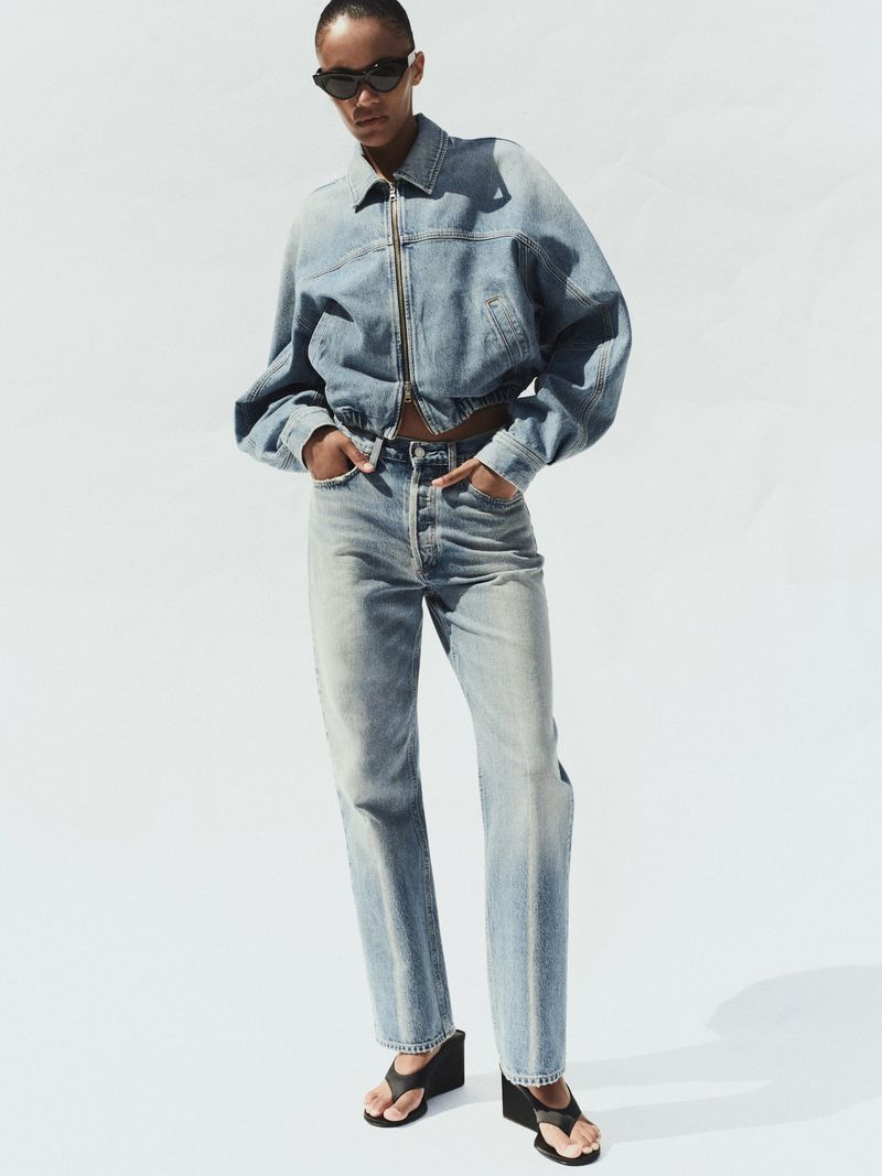 Model wearing a matching denim jacket and jeans with black sunglasses against a white background