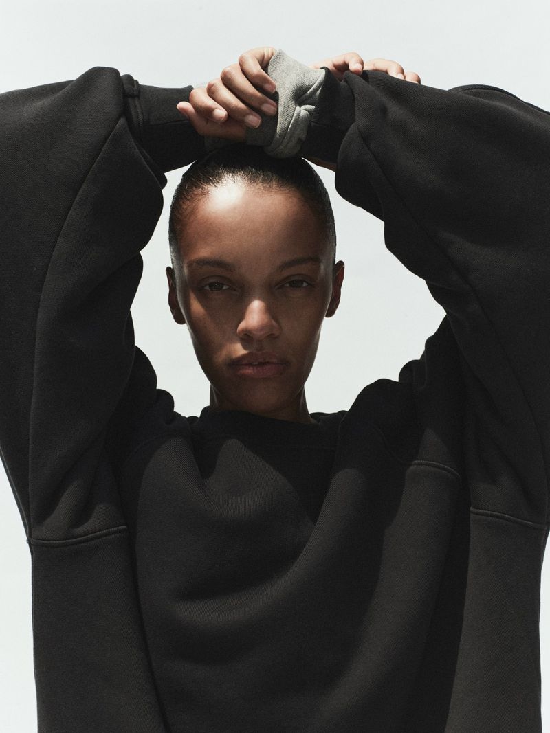 Model Kukua Williams wearing a black sweatshirt with hands clasped behind her head against a plain light background for AGOLDE Spring 2026.
