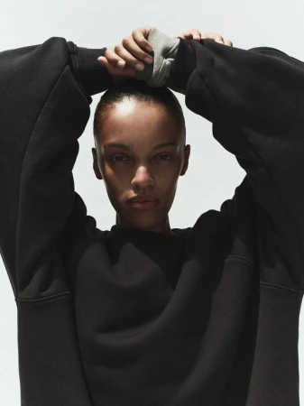 Model Kukua Williams wearing a black sweatshirt with hands clasped behind her head against a plain light background for AGOLDE Spring 2026.