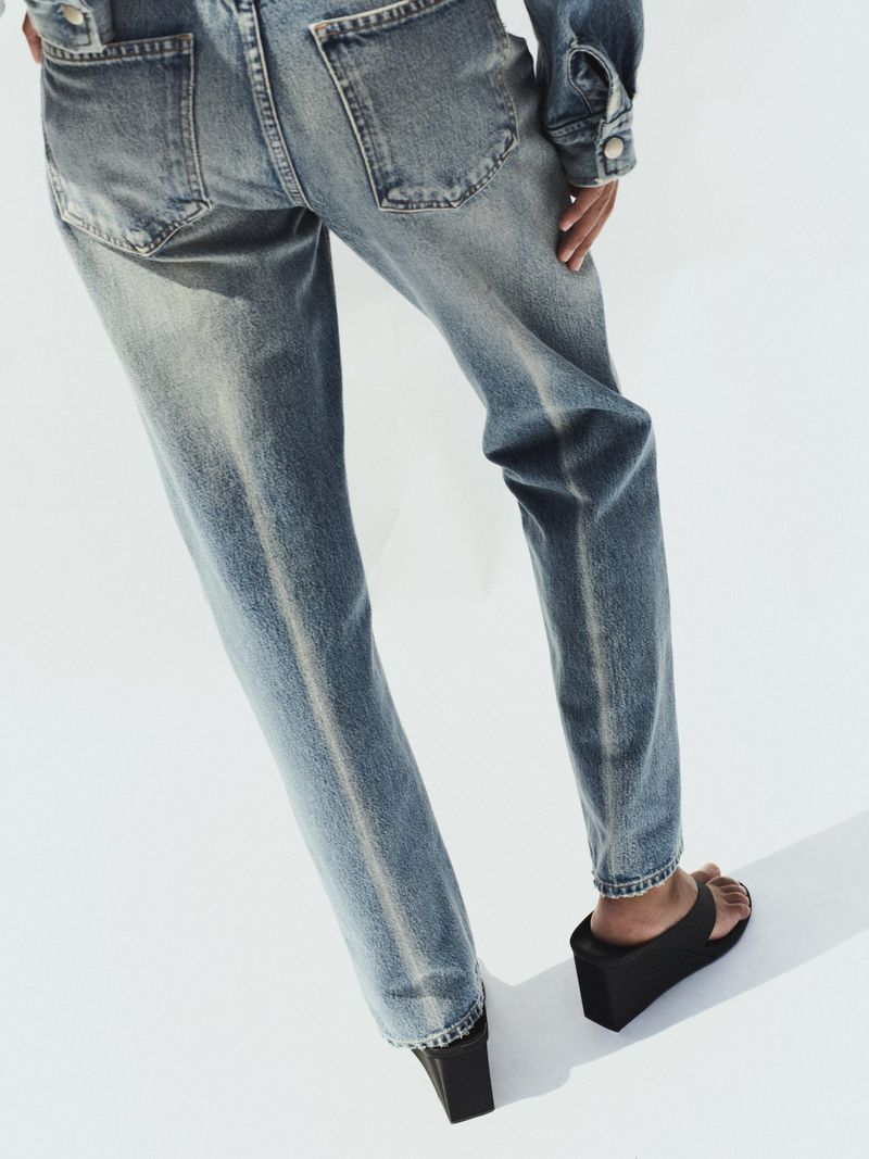 Rear view of a model wearing distressed blue denim jeans and matching denim jacket standing against a white backdrop.