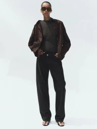 Model wearing dark sunglasses, brown button-down shirt, and black jeans against a white background.