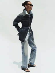 Model wearing a dark denim jacket and light wash jeans standing against a plain white background for AGOLDE.