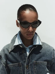 Model wearing dark sunglasses, a light blue collared shirt, and a denim jacket against a bright white background.