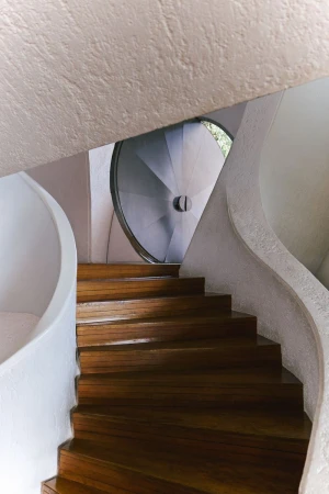 Wooden staircase curving upward between white textured walls toward a circular window for Literary Sport SS 2026