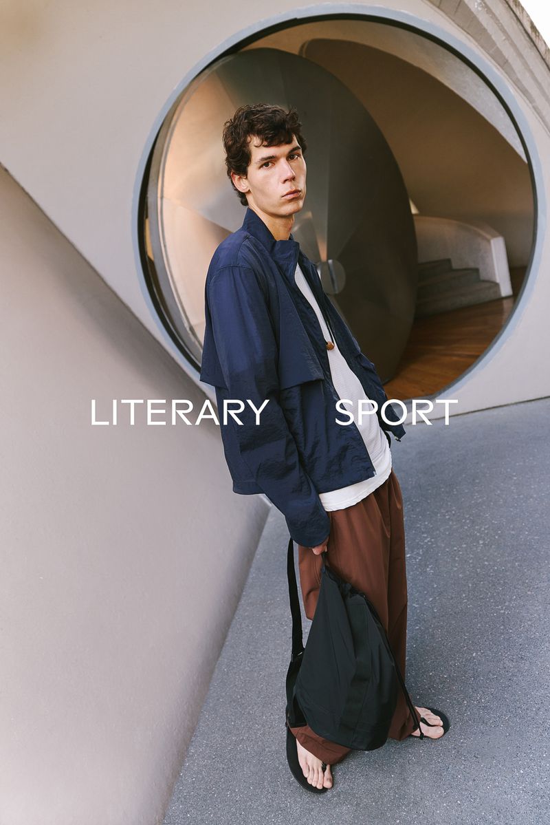Model in a navy blue jacket and brown trousers leaning against a curved concrete wall with a large circular window.