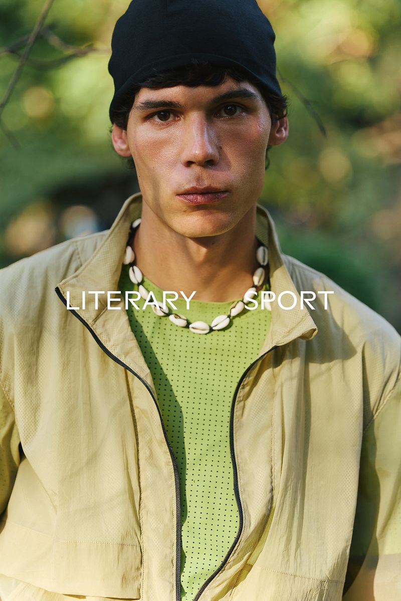 Close-up portrait of a male model wearing a black beanie, shell necklace, lime green shirt, and a light-colored jacket outdoors.