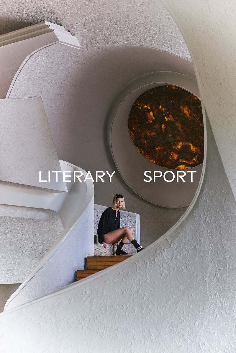 Model seated on a wooden staircase inside an architectural interior with curved white walls and a glowing circular window.