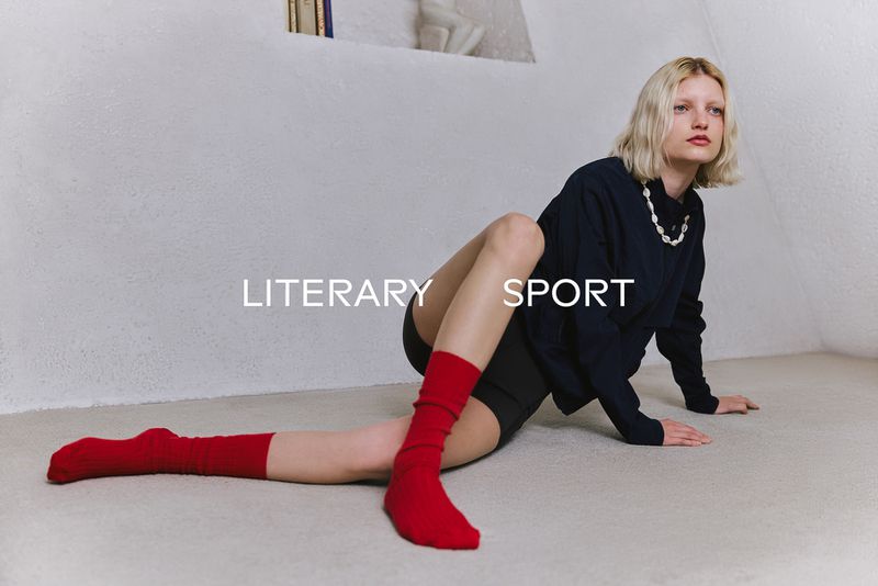 Model seated on a pale floor wearing a dark top, black shorts, and bright red socks against a textured white wall.