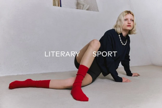 Model seated on a pale floor wearing a dark top, black shorts, and bright red socks against a textured white wall.