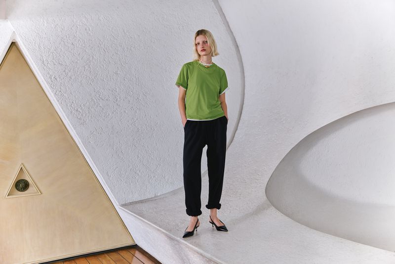 Model wearing a green t-shirt and black trousers posing inside an architectural interior with textured white curved walls.
