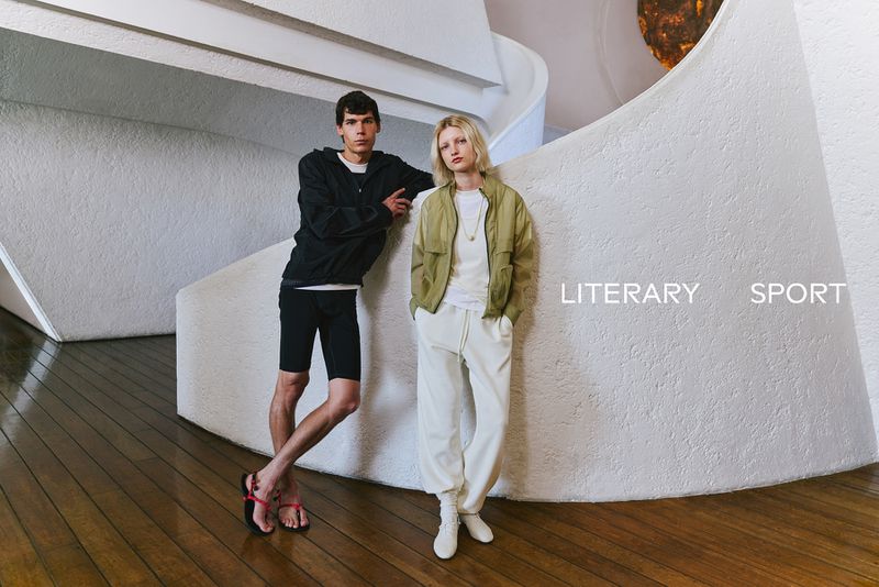 Two models posing against a curved white textured wall inside a modern building with dark wooden floors for LITERARY SPORT SS 2026