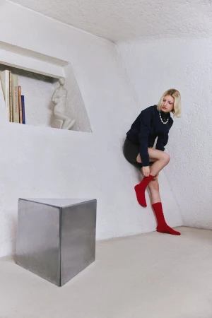 Model wearing red socks and a dark top leaning against a white textured wall next to a metal block sculpture.