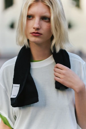 Model with blonde hair and a black towel around her neck wearing a white t shirt against an outdoor street background