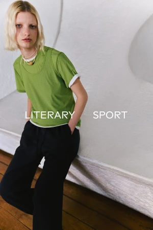 Model in green mesh t-shirt and black trousers posing against a textured white wall for Literary Sport SS 2026.