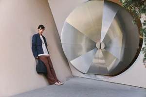 Model leaning against a wall beside a large metallic circular architectural feature for LITERARY SPORT SS 2026