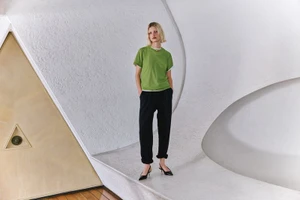 Model wearing a green t-shirt and black trousers posing inside an architectural interior with textured white curved walls.