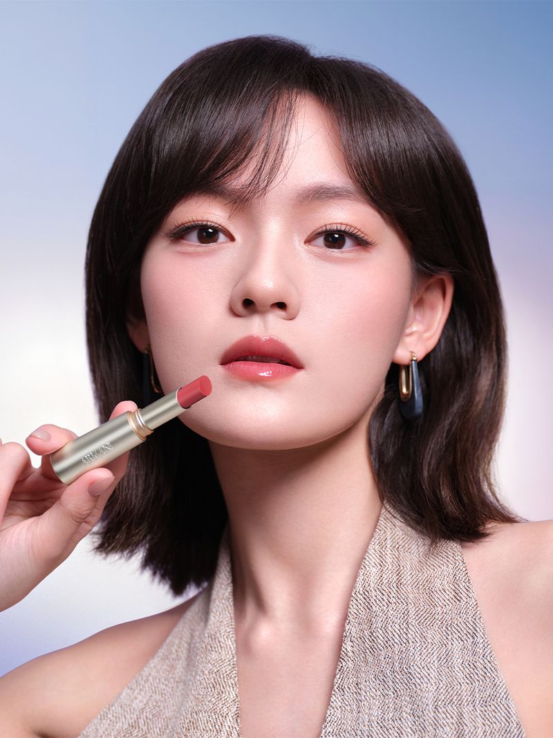 Close-up of a woman with dark hair holding a coral lipstick, posing against a gradient blue and white background for beauty shot.