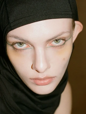 Close-up portrait of a person with pale skin, striking green eye makeup, and wearing a black head covering looking intensely at the camera.