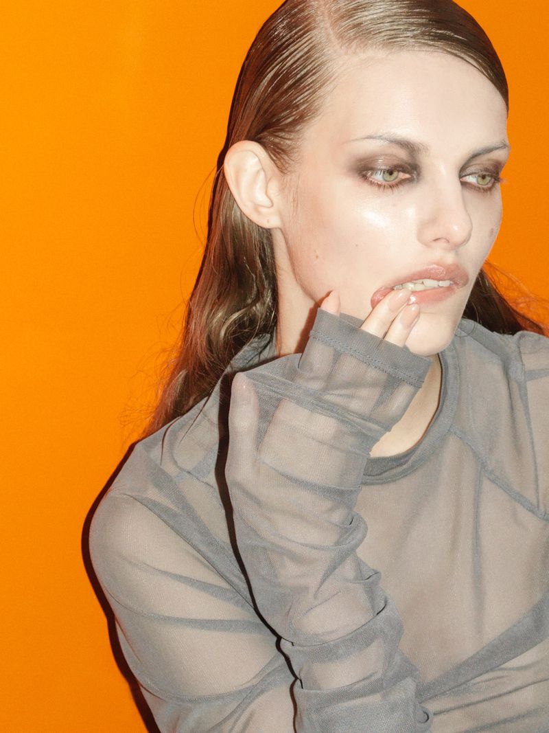 Close-up studio portrait of an adult female fashion model with smokey eye makeup, sheer top, and wet-look hairstyle against an orange backdrop.