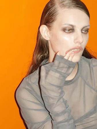 Close-up studio portrait of an adult female fashion model with smokey eye makeup, sheer top, and wet-look hairstyle against an orange backdrop.