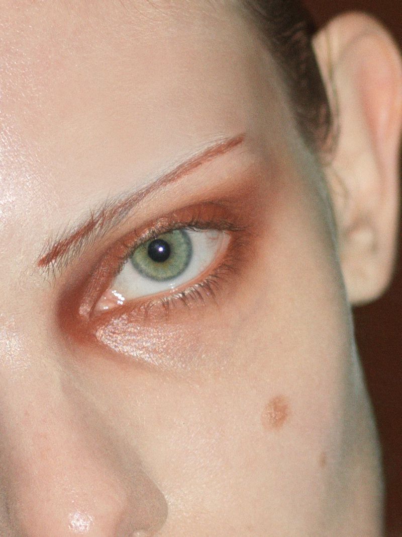 Close-up shot of a woman's face highlighting green eye with brown eye shadow, brow line makeup, fair skin, and a beauty mark.