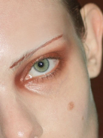 Close-up shot of a woman's face highlighting green eye with brown eye shadow, brow line makeup, fair skin, and a beauty mark.