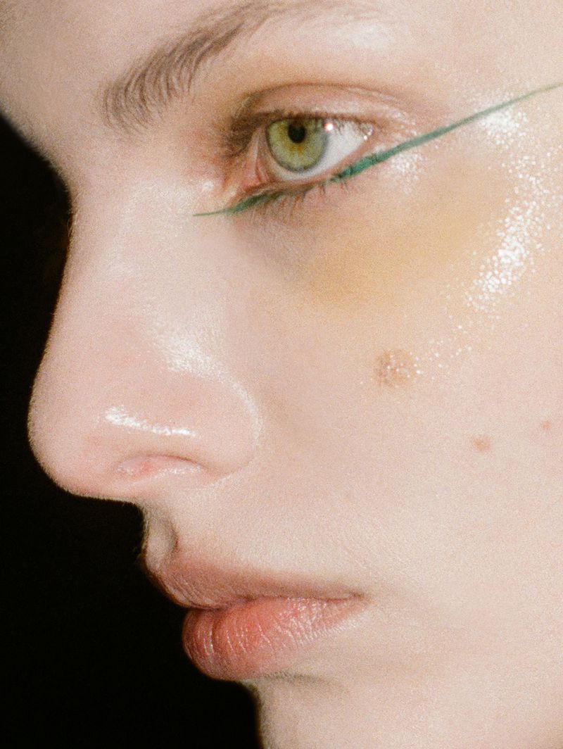 Close-up portrait of an adult female face with pale skin, green eyes, and green eyeliner, set against a dark background.