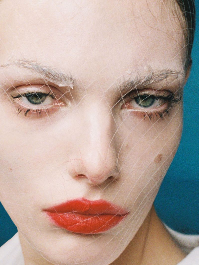 Close-up of a woman's face with red lipstick, white eyebrows and a white net over her face against a blue background.