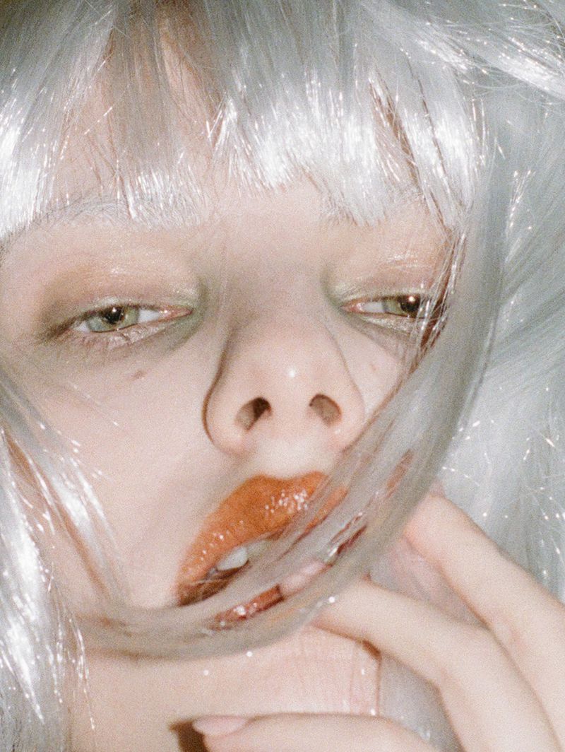 Close-up portrait of a woman with fair skin and white hair, featuring shimmering eyeshadow and glossy lips, set indoors.