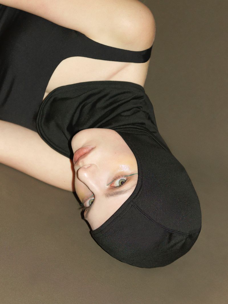 Woman with fair skin poses in a black hooded top against a brown background, looking up with neutral facial expression in studio lighting.