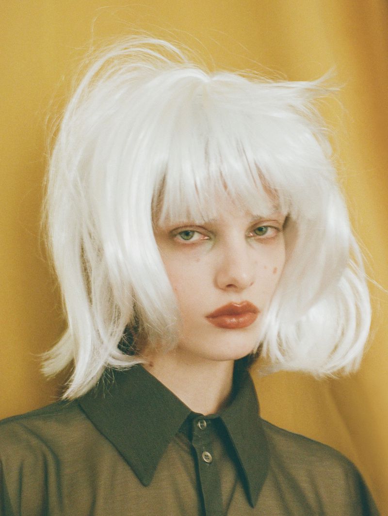 Close-up studio portrait of a young woman with white wig and olive shirt against a golden yellow draped background.