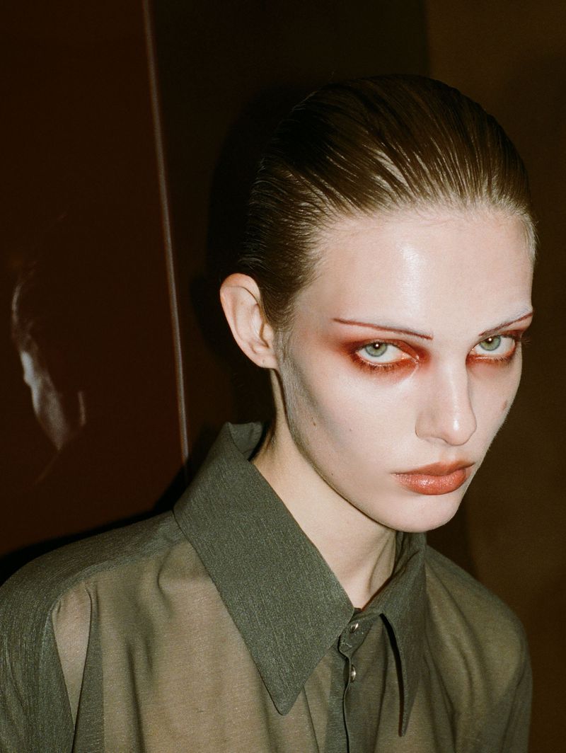 Close-up portrait of a young adult female fashion model with slicked-back hair, wearing a sheer button-down olive-green shirt.