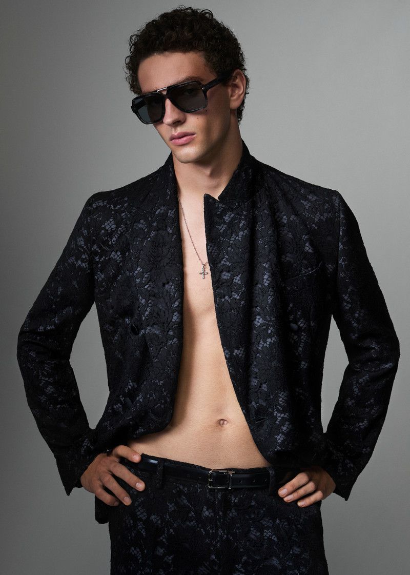 Man with sunglasses in black lace patterned suit, silver cross necklace posing in front of gray studio backdrop for fashion editorial.