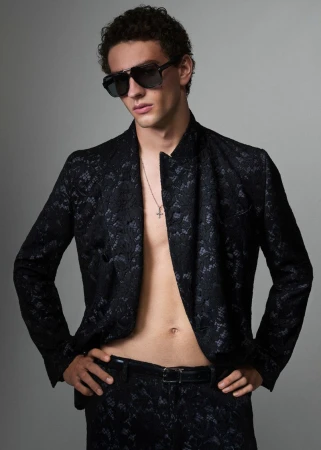 Man with sunglasses in black lace patterned suit, silver cross necklace posing in front of gray studio backdrop for fashion editorial.