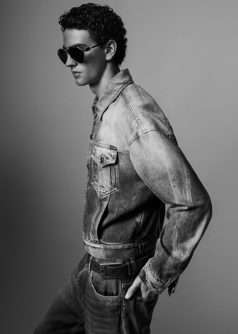 Black and white studio shot of a stylish adult male model wearing sunglasses, a denim jacket, and jeans posing against a plain background.
