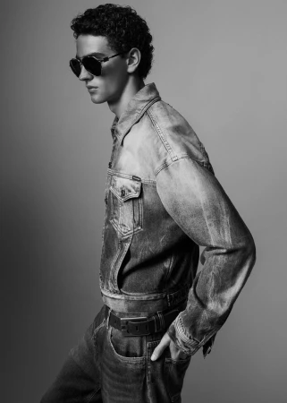Black and white studio shot of a stylish adult male model wearing sunglasses, a denim jacket, and jeans posing against a plain background.