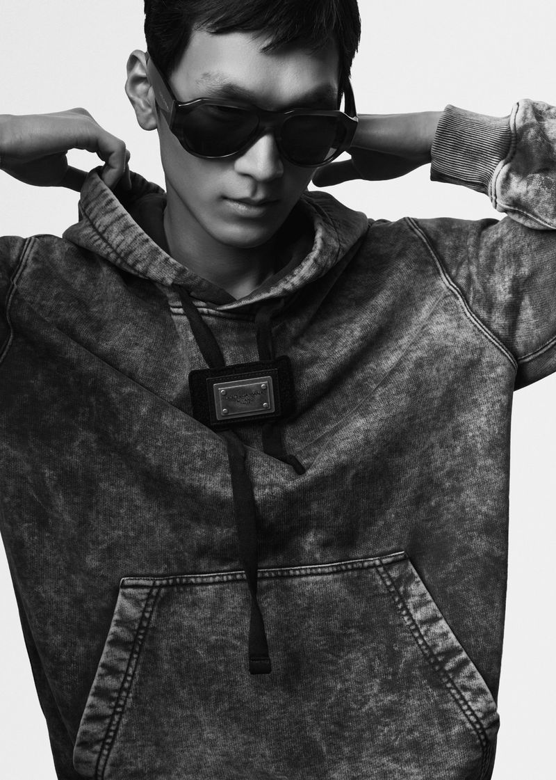 Model wearing dark sunglasses and an acid-wash hoodie posing against a solid white background in a studio portrait