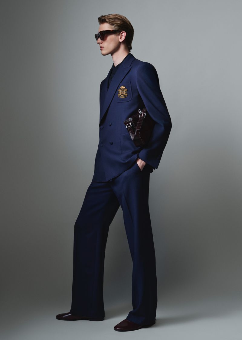 Full-length portrait of a male model wearing a navy blue double-breasted suit and sunglasses against a grey studio backdrop.