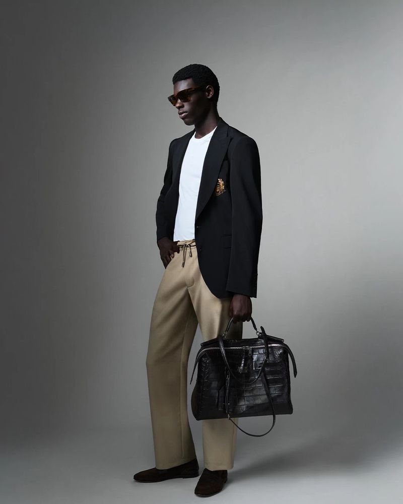 Man wearing a black blazer over a white t-shirt and tan trousers holding a large black leather bag against a grey background.