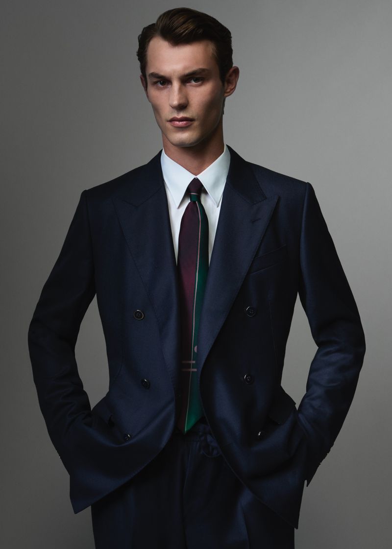 A man in a dark double-breasted suit, white shirt, and tie posing against a neutral grey studio backdrop.