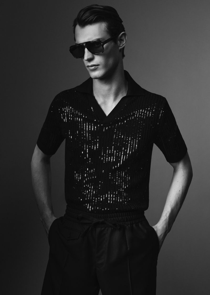 Model wearing dark sunglasses and a patterned dark polo shirt against a plain grey backdrop.