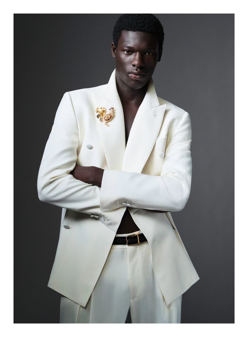 Black man wearing a white double-breasted suit jacket with a gold lapel pin and a black belt against a grey background