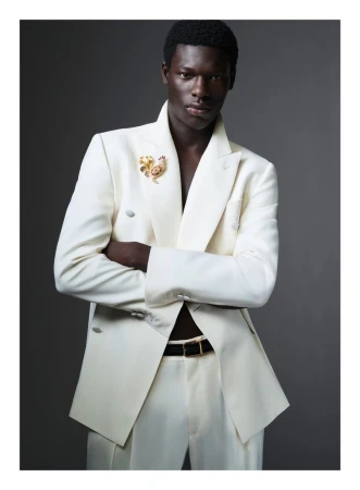 Black man wearing a white double-breasted suit jacket with a gold lapel pin and a black belt against a grey background