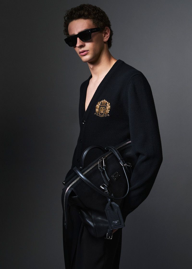 Male model in black sunglasses and a black cardigan holding a dark leather duffel bag against a grey studio backdrop.