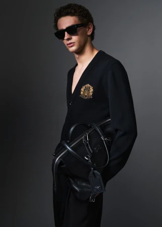 Male model in black sunglasses and a black cardigan holding a dark leather duffel bag against a grey studio backdrop.