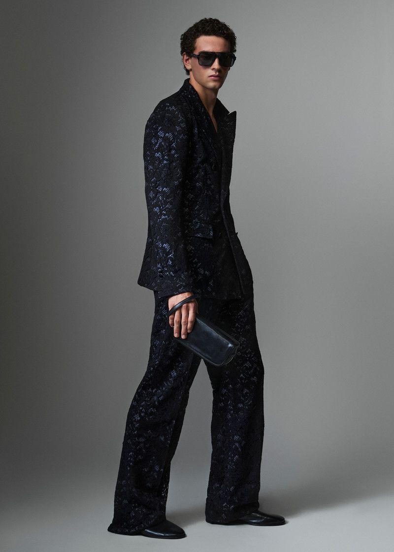 Full-length portrait of a male model wearing a dark patterned suit and sunglasses, holding a clutch against a grey studio backdrop.
