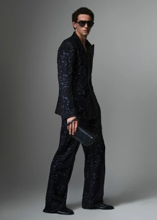 Full-length portrait of a male model wearing a dark patterned suit and sunglasses, holding a clutch against a grey studio backdrop.