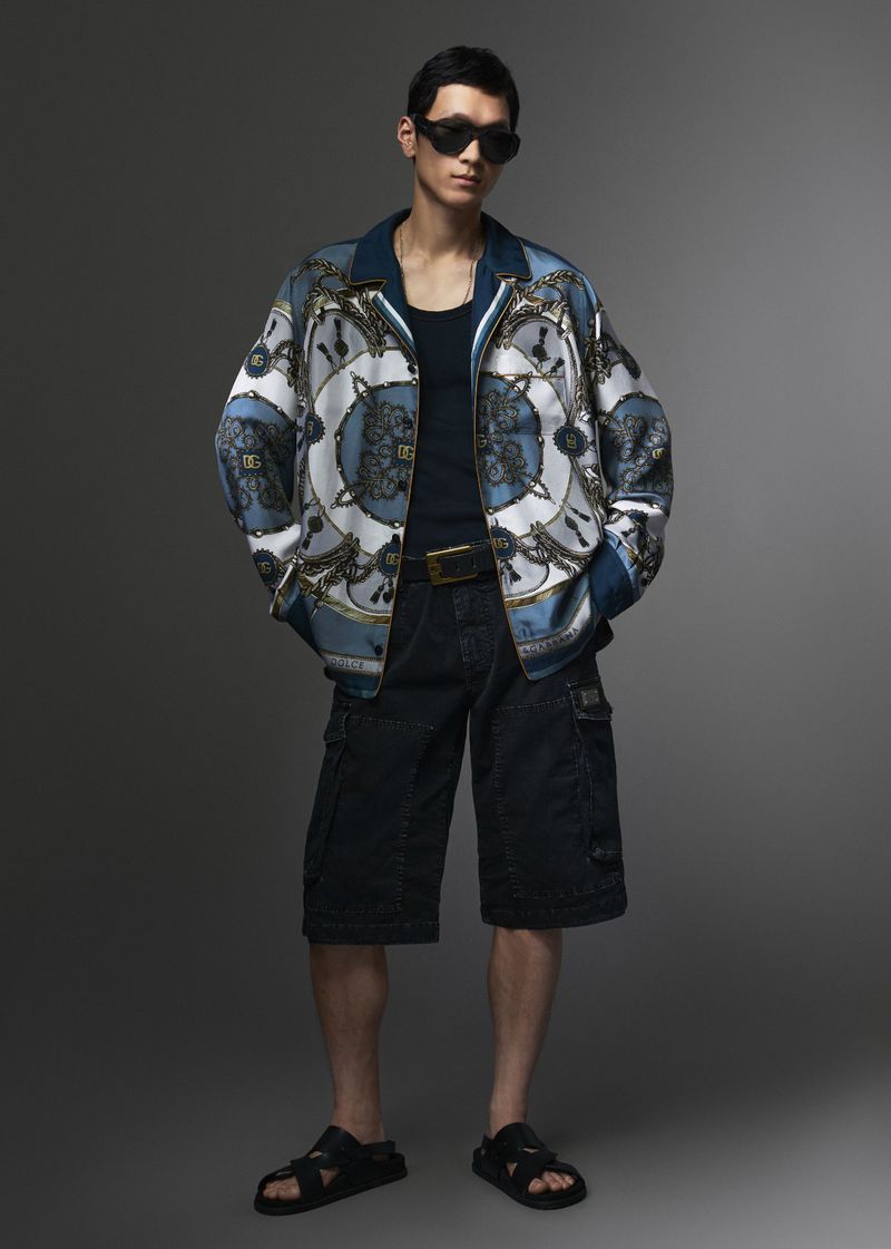 Man wearing a blue patterned silk jacket, dark tank top, cargo shorts, and sunglasses against a grey studio backdrop