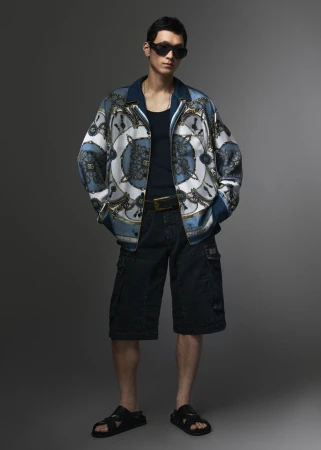 Man wearing a blue patterned silk jacket, dark tank top, cargo shorts, and sunglasses against a grey studio backdrop