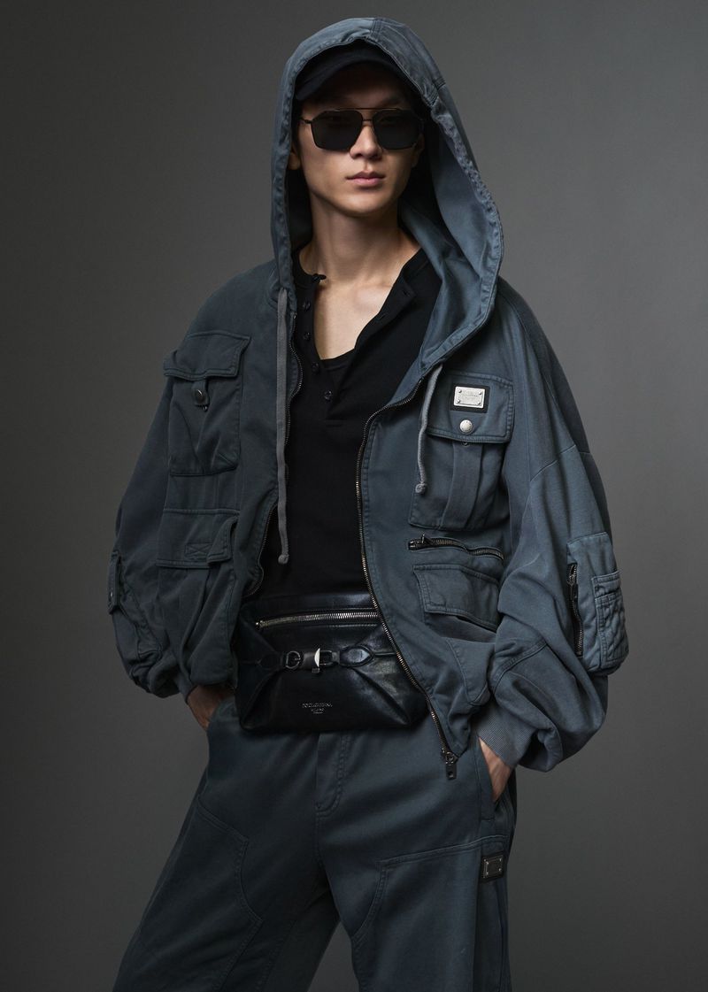 Model wearing a grey hooded utility jacket and sunglasses against a solid dark grey backdrop